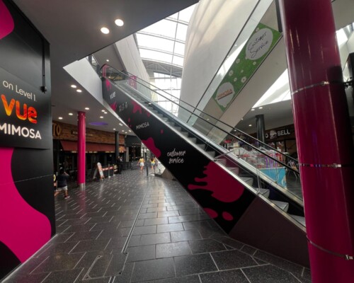 Branded escalator vinyl wrap and wayfinding graphics inside The Atrium Camberley shopping centre, featuring promotional branding and directional signage for retail and leisure destinations.
