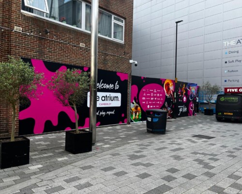 Large-format printed hoarding graphics installed along an external walkway at The Atrium Camberley shopping centre, featuring vibrant pink and black branding panels and promotional displays.